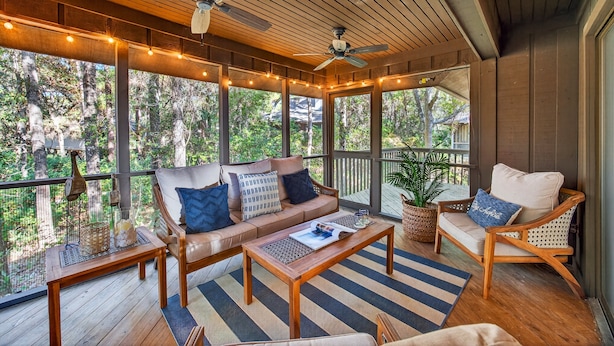 A covered outdoor seating area with wooden furniture, string lights, and a view of the forest. - Excellent Inlet Cove Cottage! Community Pool, Fishing Dock! (Johns Island)