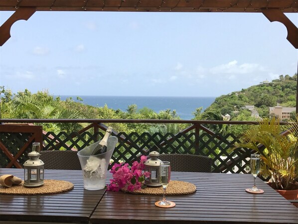 A wooden table with a view of the ocean, surrounded by greenery and a wooden pergola.