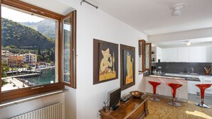 A modern kitchen with white cabinets, a black countertop, and red bar stools. A window offers a view of a harbor and buildings. - BEUST HOUSE - ADA - Balcony overlooking lake, harbor and village (Nago-Torbole)