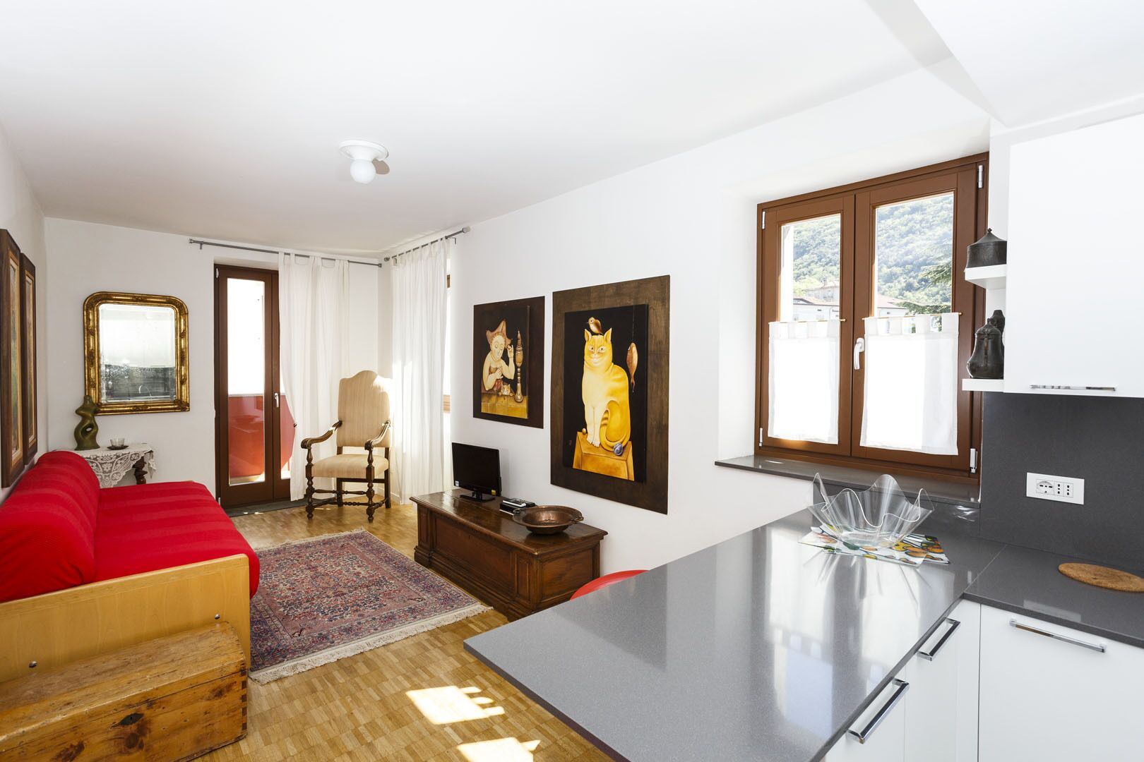 A compact living space with a red sofa, wooden chest, and a small table with a TV. There are two paintings on the wall, a chair, and a kitchen area with a window.