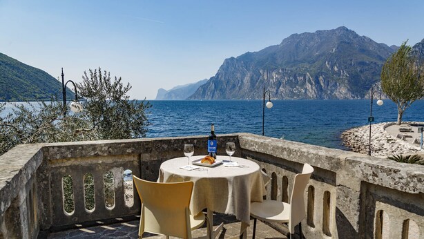 A seaside table with a bottle of wine, two glasses, and a plate with food, overlooking a mountainous coastline. - BEUST HOUSE - ADA - Balcony overlooking lake, harbor and village (Nago-Torbole)