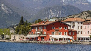 A waterfront hotel with multiple buildings, outdoor seating, and a view of mountains. - BEUST HOUSE - ADA - Balcony overlooking lake, harbor and village (Nago-Torbole)