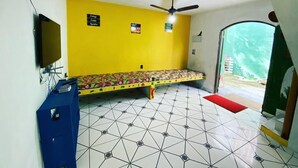 Una habitación con pared amarilla, una cama con colcha estampada, un ventilador de techo y un televisor a la izquierda.