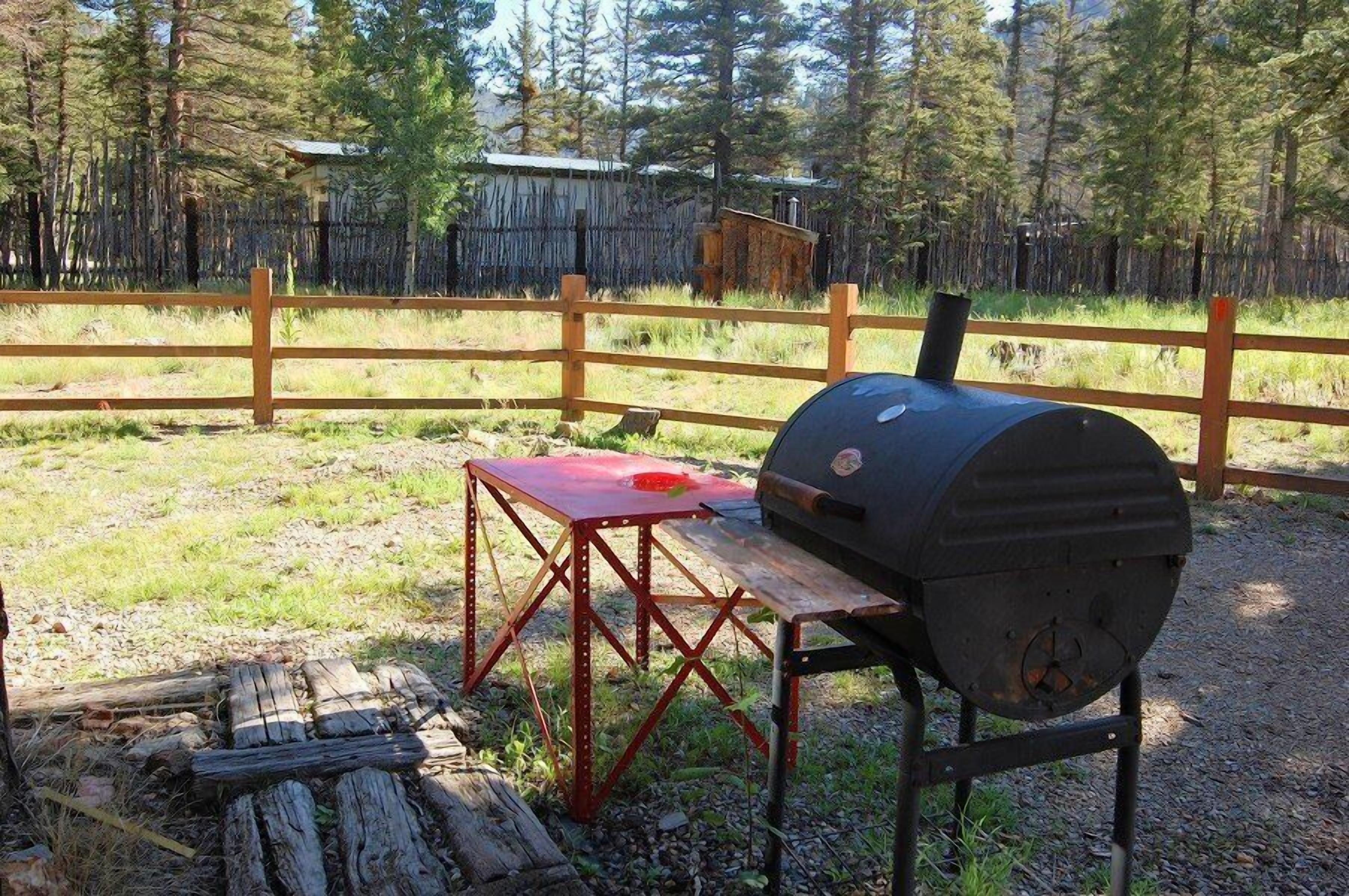 A portable barbecue grill with a metal table and benches, set in a grassy outdoor area with a wooden fence and trees in the background.