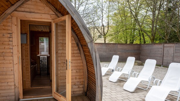Una cabaña de madera con la puerta abierta que da a un espacio para sauna y un patio con sillones blancos.