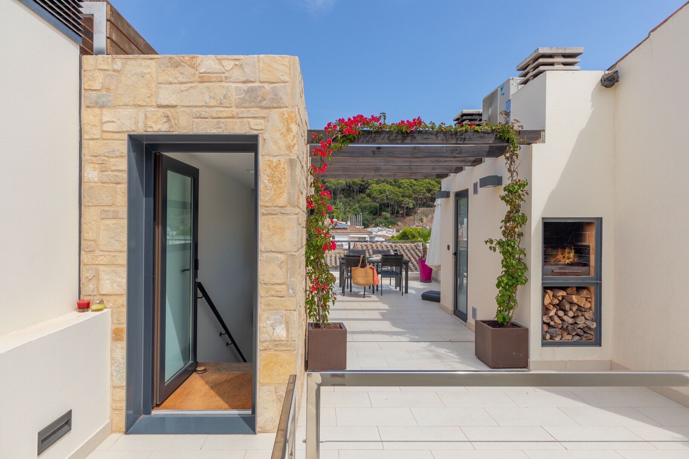 A narrow outdoor corridor with stone walls, a glass door, a wooden pergola with red flowers, and a wood-burning stove.