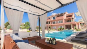 A poolside lounge area with wooden benches and a view of a swimming pool and a pink building in the background. - Apartment in Grebaštica with Seaview, Terrace, Air condition, WIFI (886-3) (Grebaštica)
