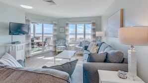 A spacious living room with a balcony, a flat-screen TV, and a large window offering a view of the beach. - Port O'Call B 203, Direct Ocean View! (Isle Of Palms)