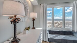 A room with a large window offering a view of the beach, a white console table with two lamps, and a gray sofa. - Port O'Call B 203, Direct Ocean View! (Isle Of Palms)