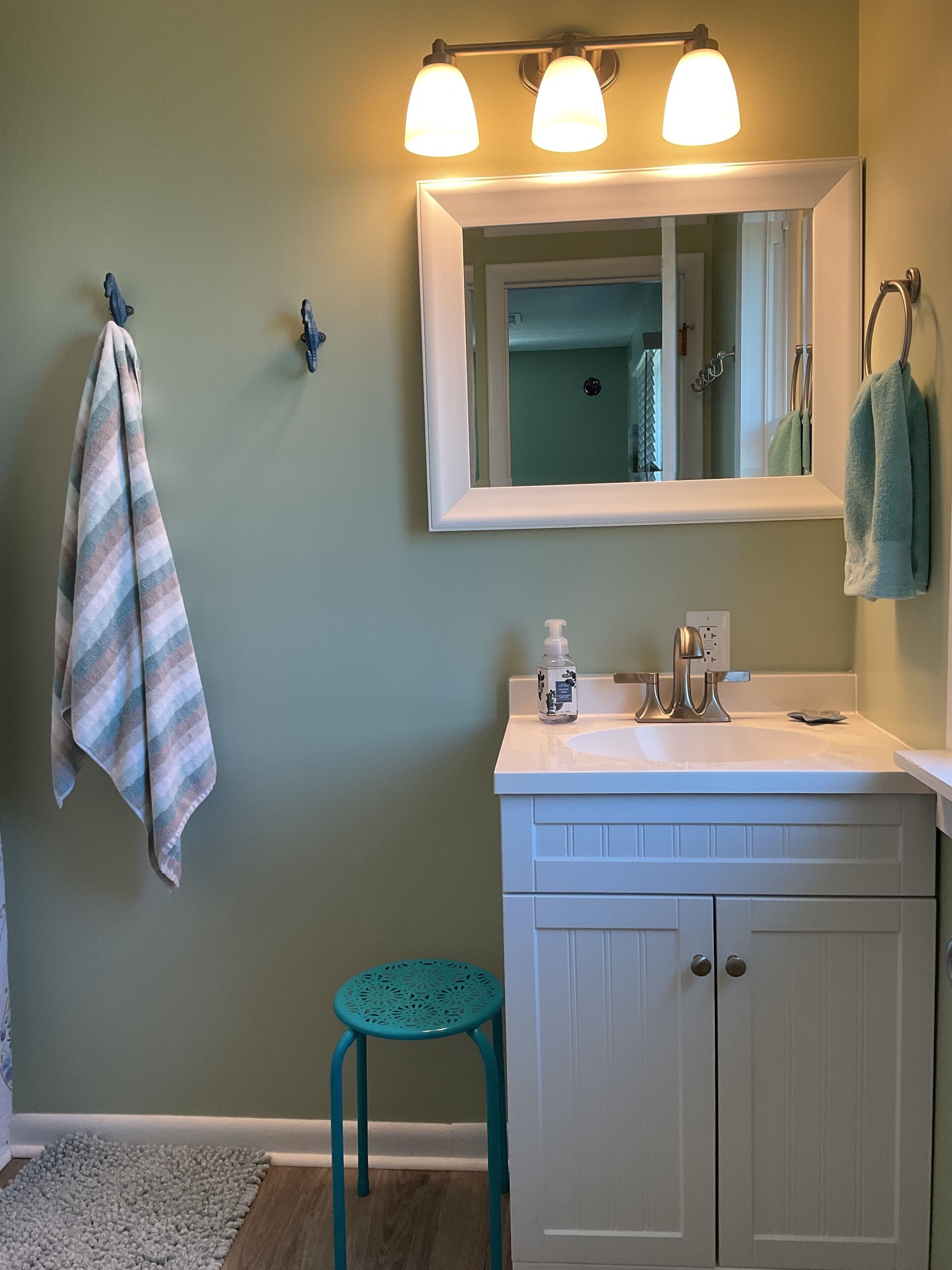 A bathroom with a white vanity, a mirror, and a sink. There is a towel rack with a striped towel and a teal towel. A small teal stool is next to the vanity.