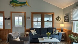 Un salon chaleureux avec un canapé bleu, un fauteuil gris et une table basse. Un grand décor représentant un poisson orne le mur, ainsi qu’une boussole et une lampe sur la table d’appoint.
