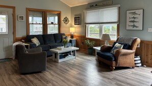 Un salon chaleureux avec un canapé bleu foncé, un fauteuil en osier et une table basse. On y trouve une fenêtre avec des stores, une lampe et une étoile de mer décorative accrochée au mur.