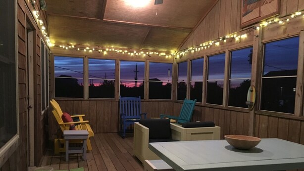 Un espace extérieur abrité, doté de bancs et de chaises en bois, de guirlandes lumineuses et de grandes fenêtres offrant une vue sur le ciel du soir.