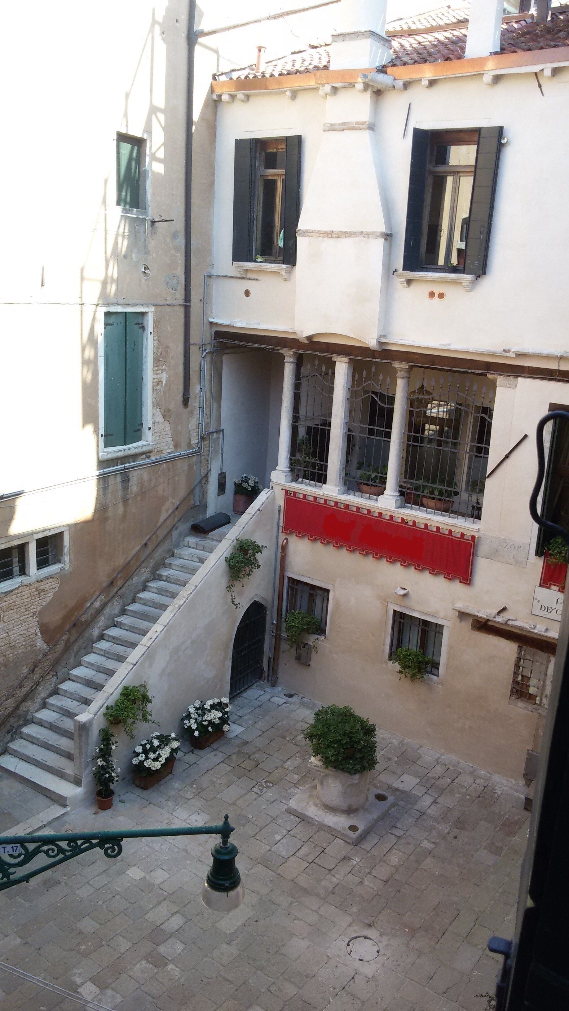 A narrow courtyard with a staircase leading to an upper floor, surrounded by buildings with balconies and potted plants.