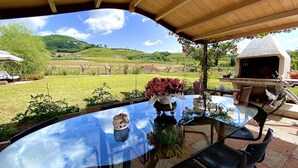 Uma área de refeições ao ar livre com mesa de vidro, cadeiras, churrasqueira e vista para um vinhedo.