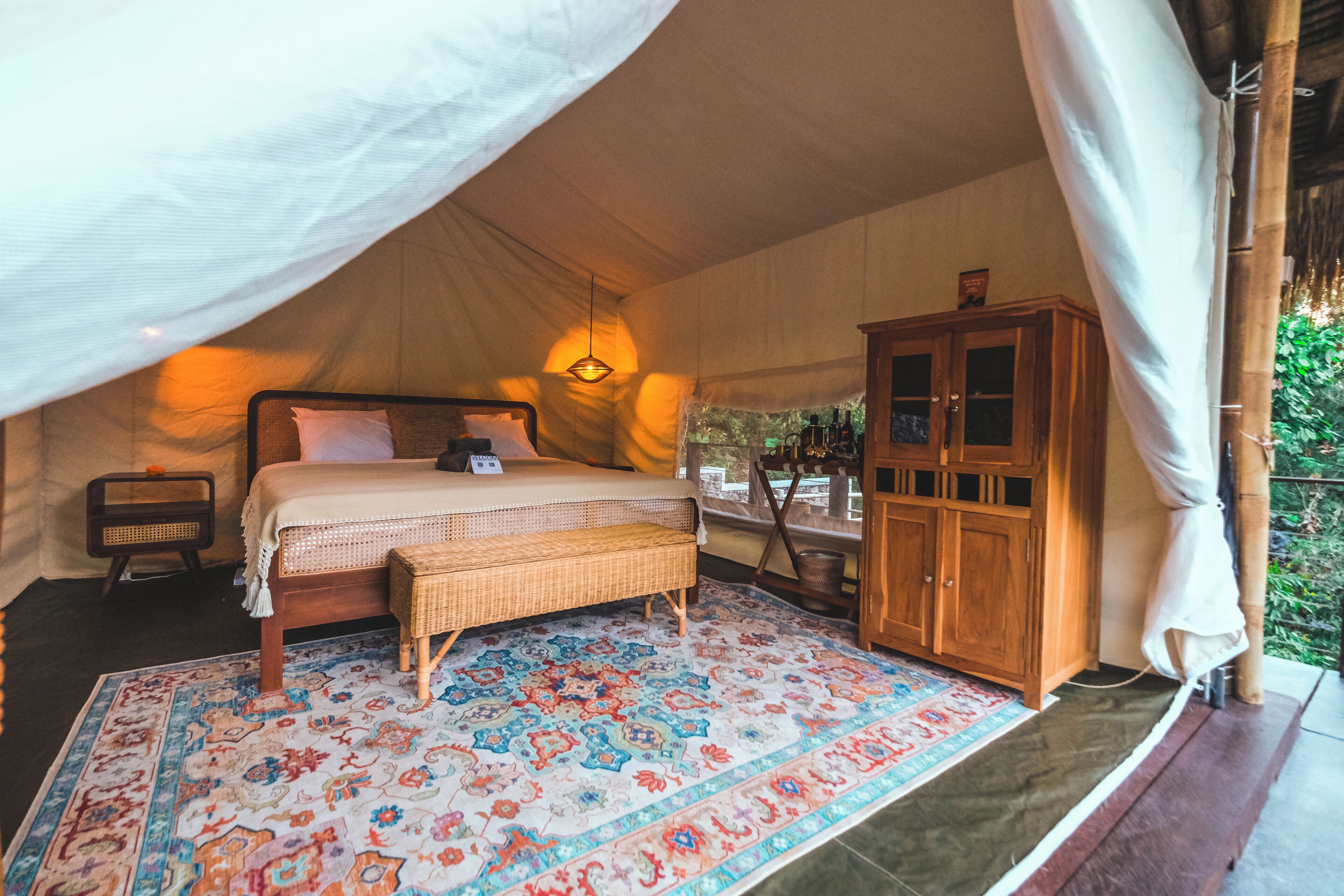 A tented room with a bed, a wooden cabinet, a wicker bench, and a patterned rug.