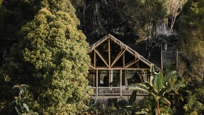 A wooden cabin with a triangular roof, surrounded by dense forest.
