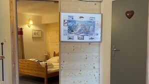 A bedroom with a wooden headboard, a bed, a nightstand, a red stool, a door with a heart decoration, and a wall with a framed picture and a map.