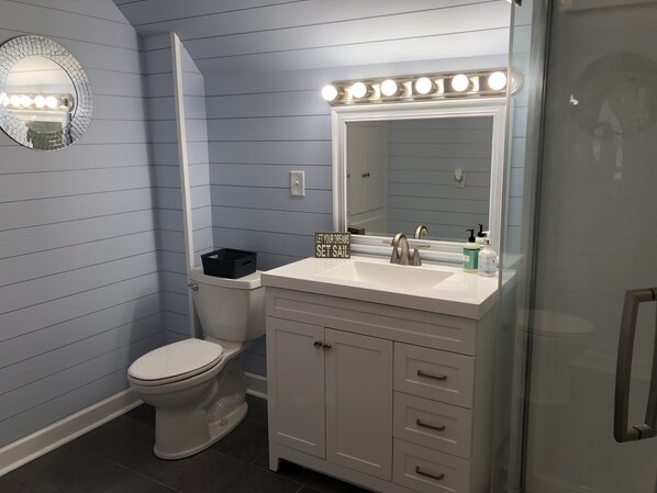 Une salle de bain avec un lavabo blanc, un miroir et un meuble-lavabo. On y trouve des toilettes sur lesquelles est posé un panier noir. Les murs sont peints en bleu et un luminaire est installé au-dessus du miroir.