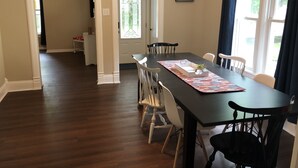 Un comedor con una mesa de madera oscura, sillas blancas y un corredor estampado. Hay una ventana con cortinas y una puerta que conduce a otra habitación.