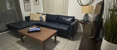 A living room with a dark leather sofa, a wooden coffee table, a TV stand with a flat-screen TV, a lamp, and a potted plant.