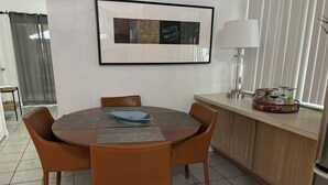 Ein Essbereich mit einem runden Tisch, vier Stühlen, einem gerahmten Bild an der Wand, einer Lampe auf der Arbeitsfläche und einer Küchenzeile mit Schränken und Spüle.