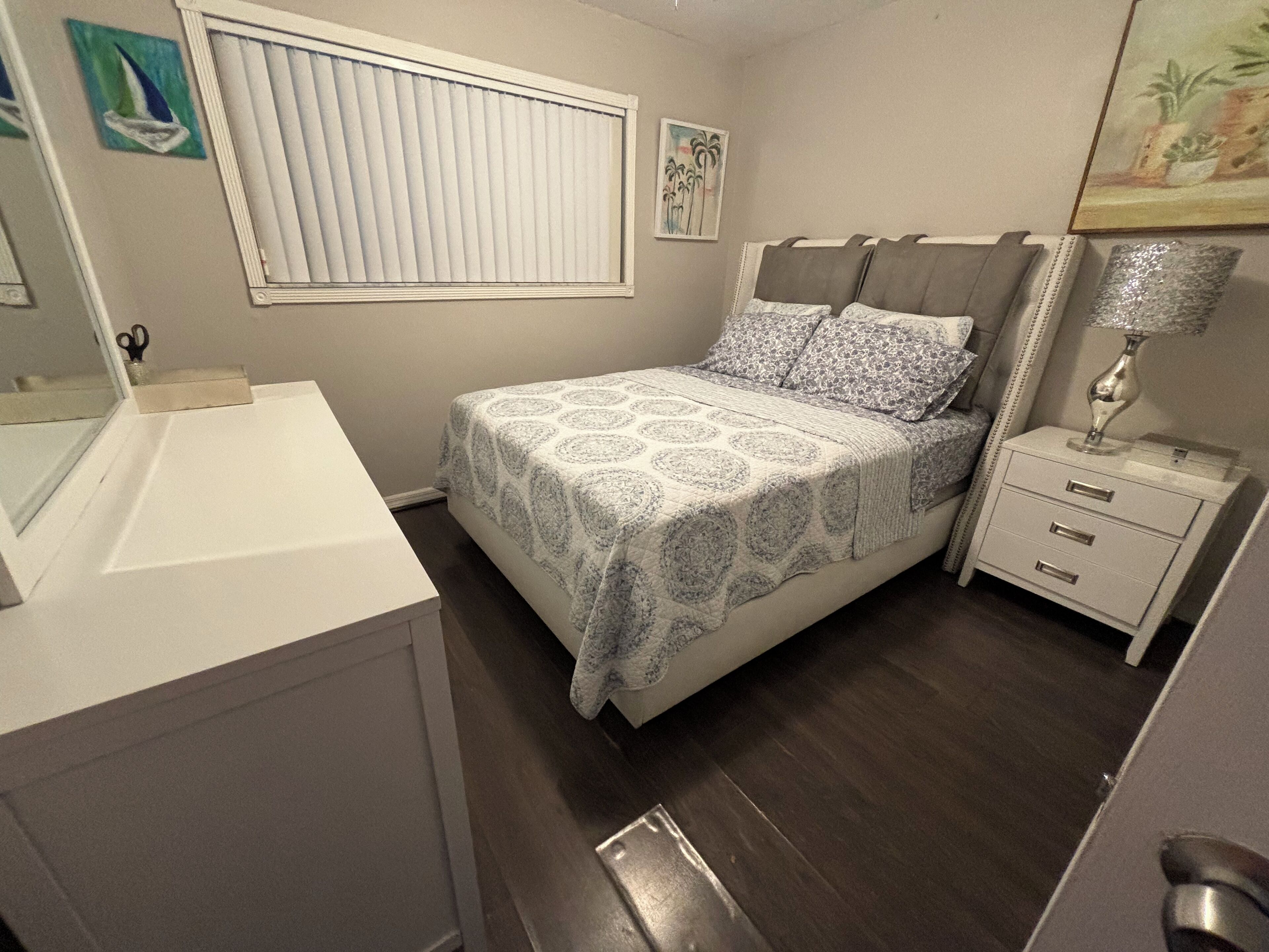 A bedroom with a bed, a nightstand, a lamp, a window with blinds, and a painting on the wall.