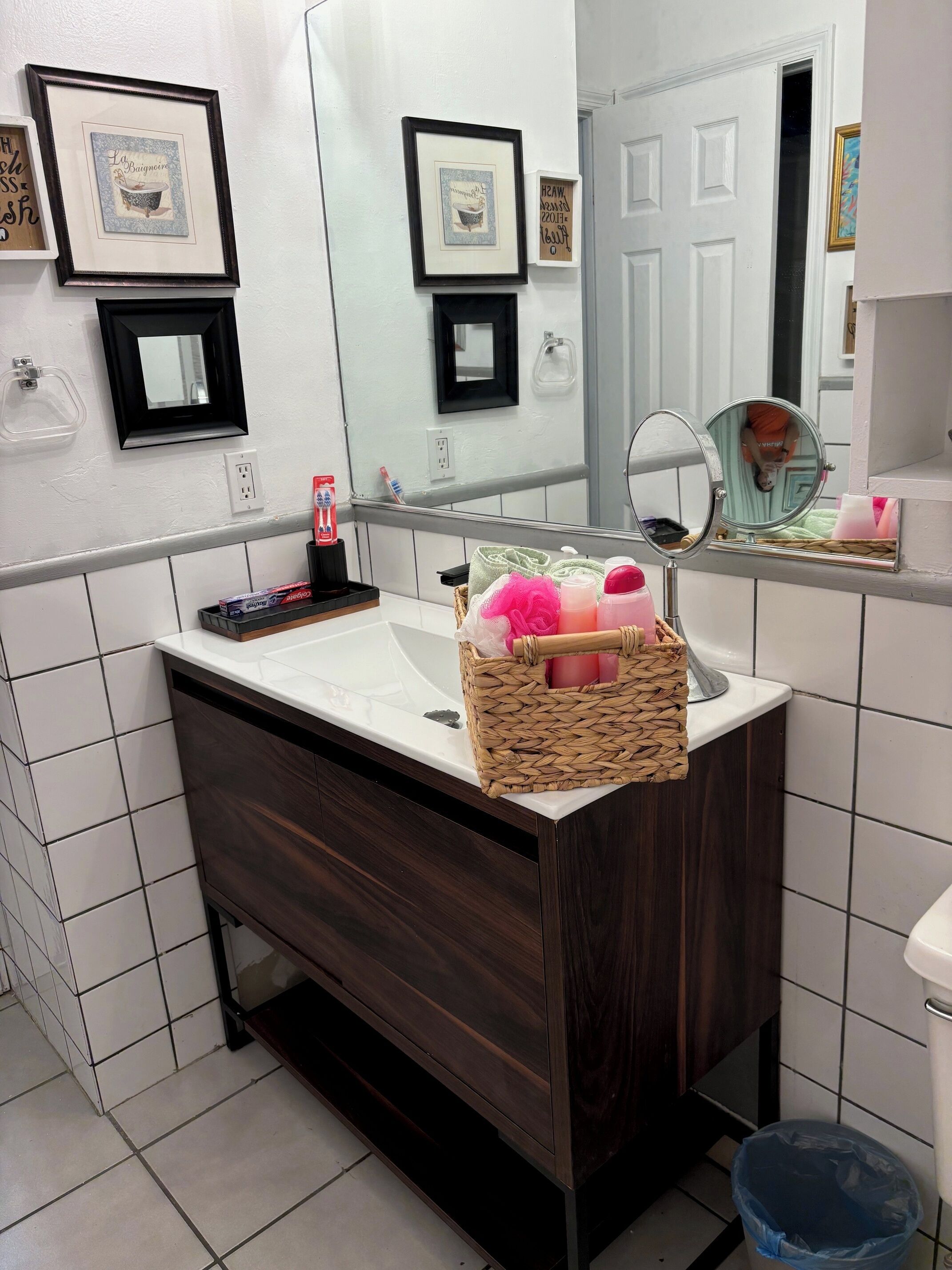 Un baño con tocador de madera oscura, azulejos blancos, un espejo y una cesta de artículos de aseo.