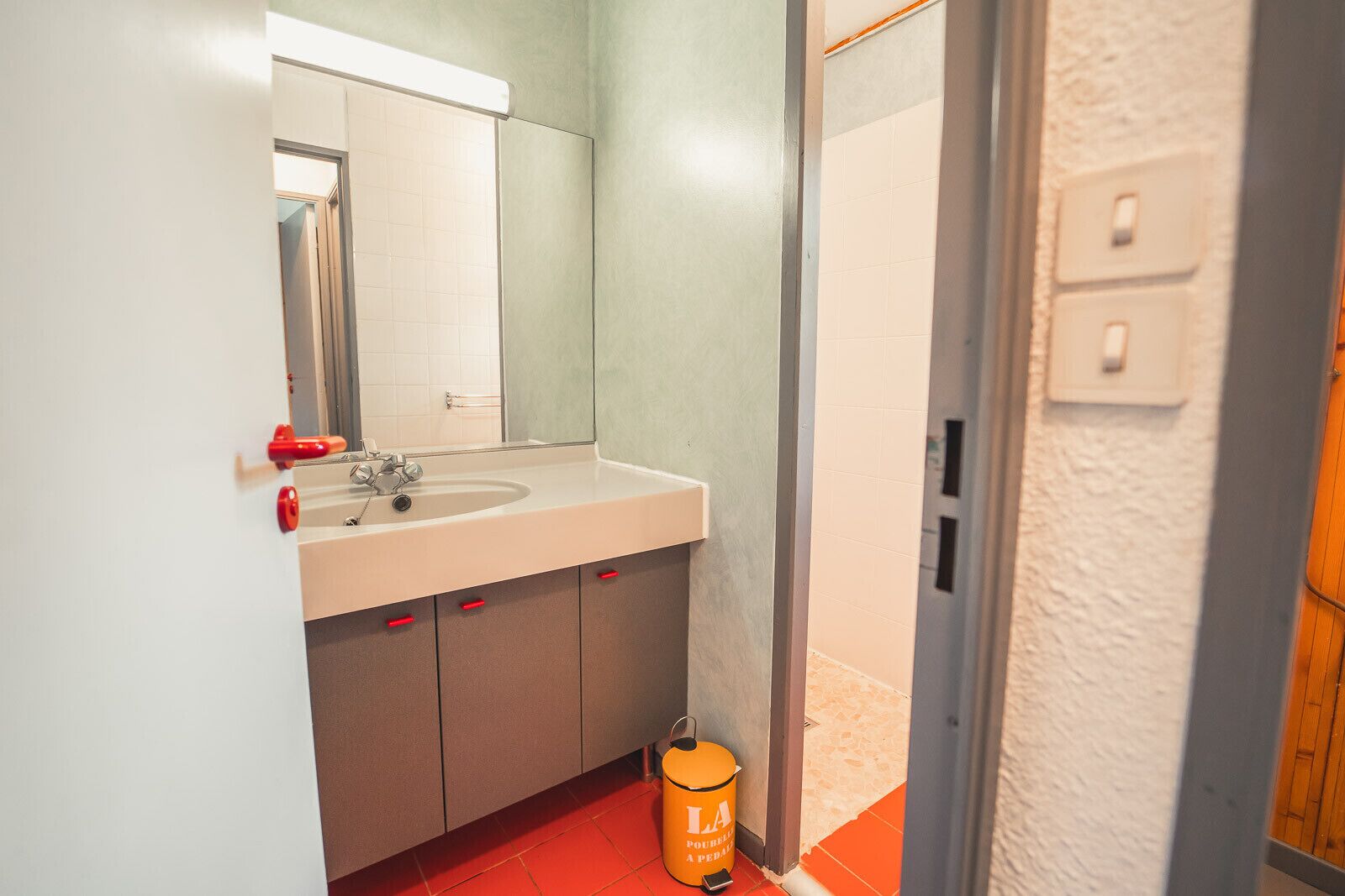 A bathroom with a sink, mirror, and a red bucket labeled 'LA POUPEAU'.
