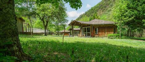 A rustic wooden cabin with a sloped roof, surrounded by lush greenery and trees.