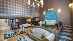 Une chambre d’hôtel moderne dotée d’un grand lit, d’un coin salon avec un canapé et une table à manger, d’un lustre et d’un grand miroir qui reflète la pièce.