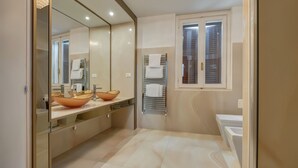 Une salle de bain moderne avec un grand miroir, deux vasques, dont une en forme de grand bol, un porte-serviettes et une fenêtre avec des stores.