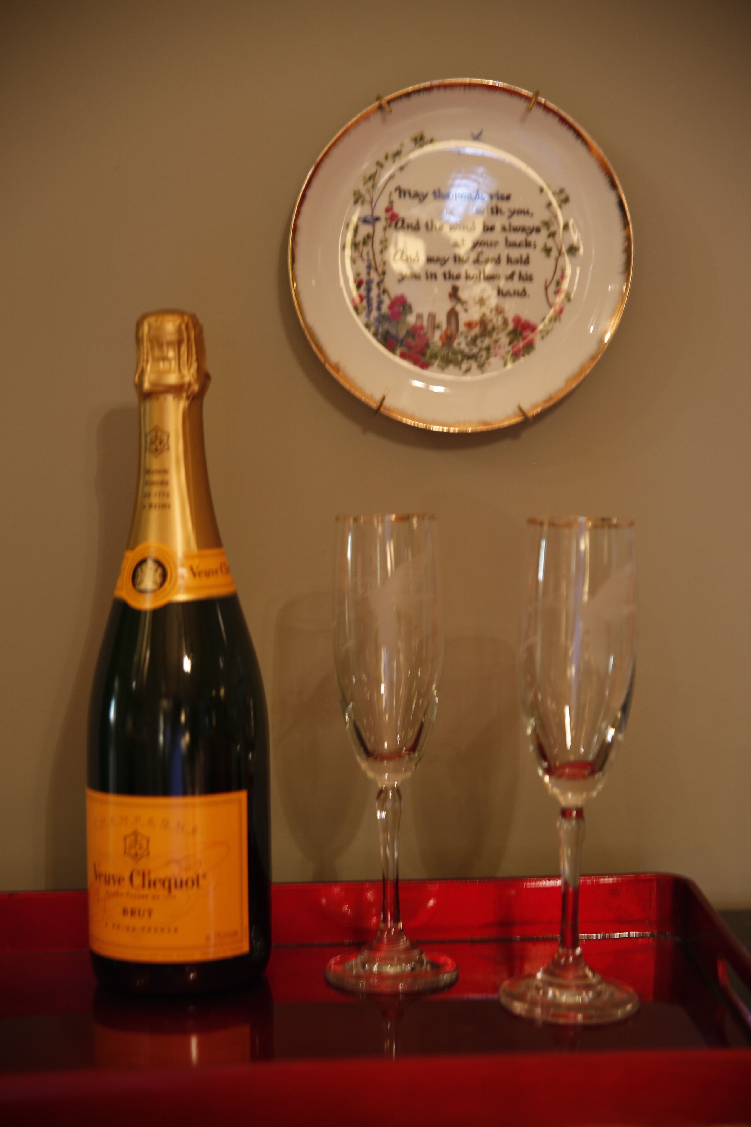 Een fles champagne, twee lege glazen en een decoratief bord met een citaat op een rode schaal.