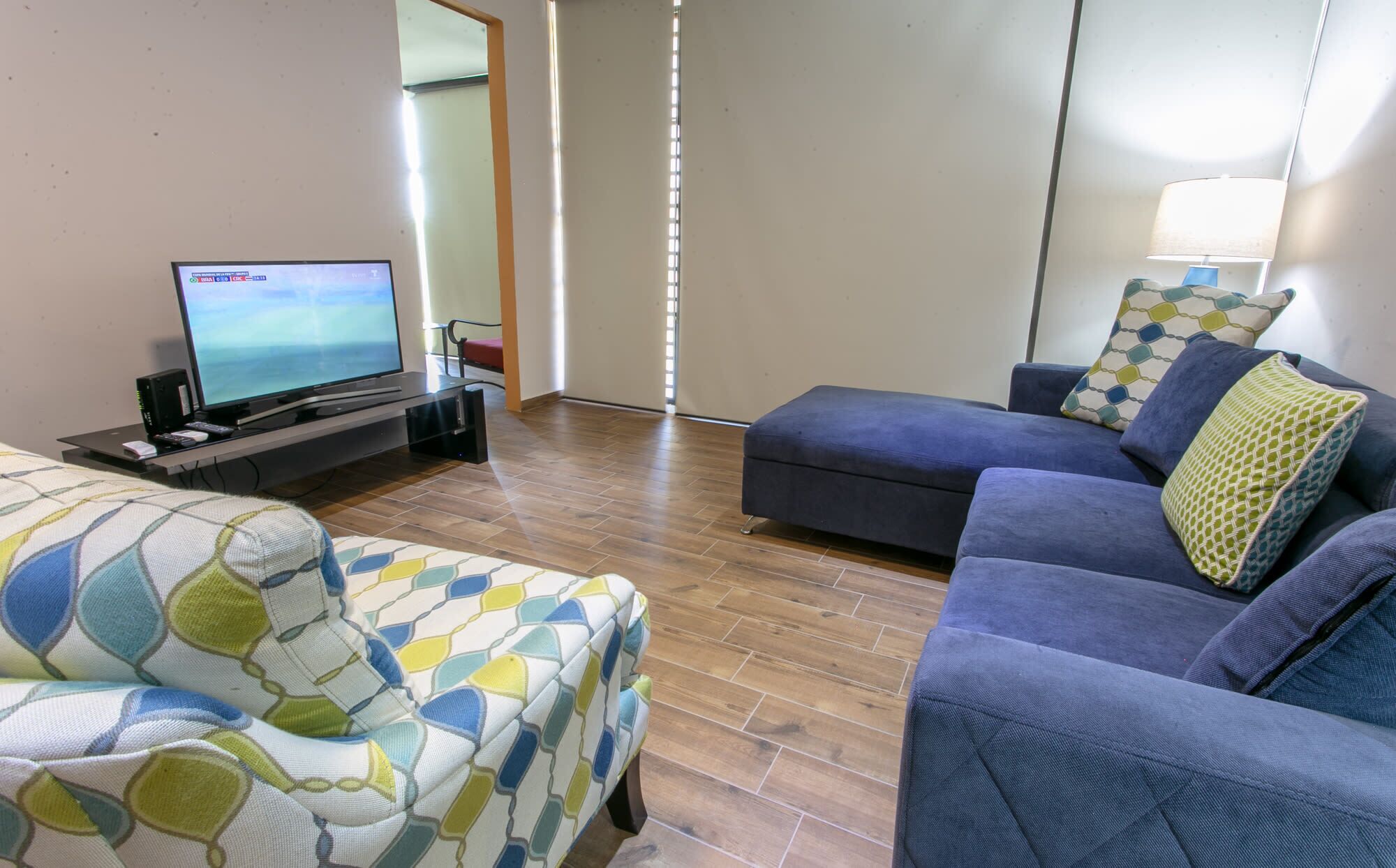 A living room with a flat-screen TV, a blue sofa, and a patterned armchair.
