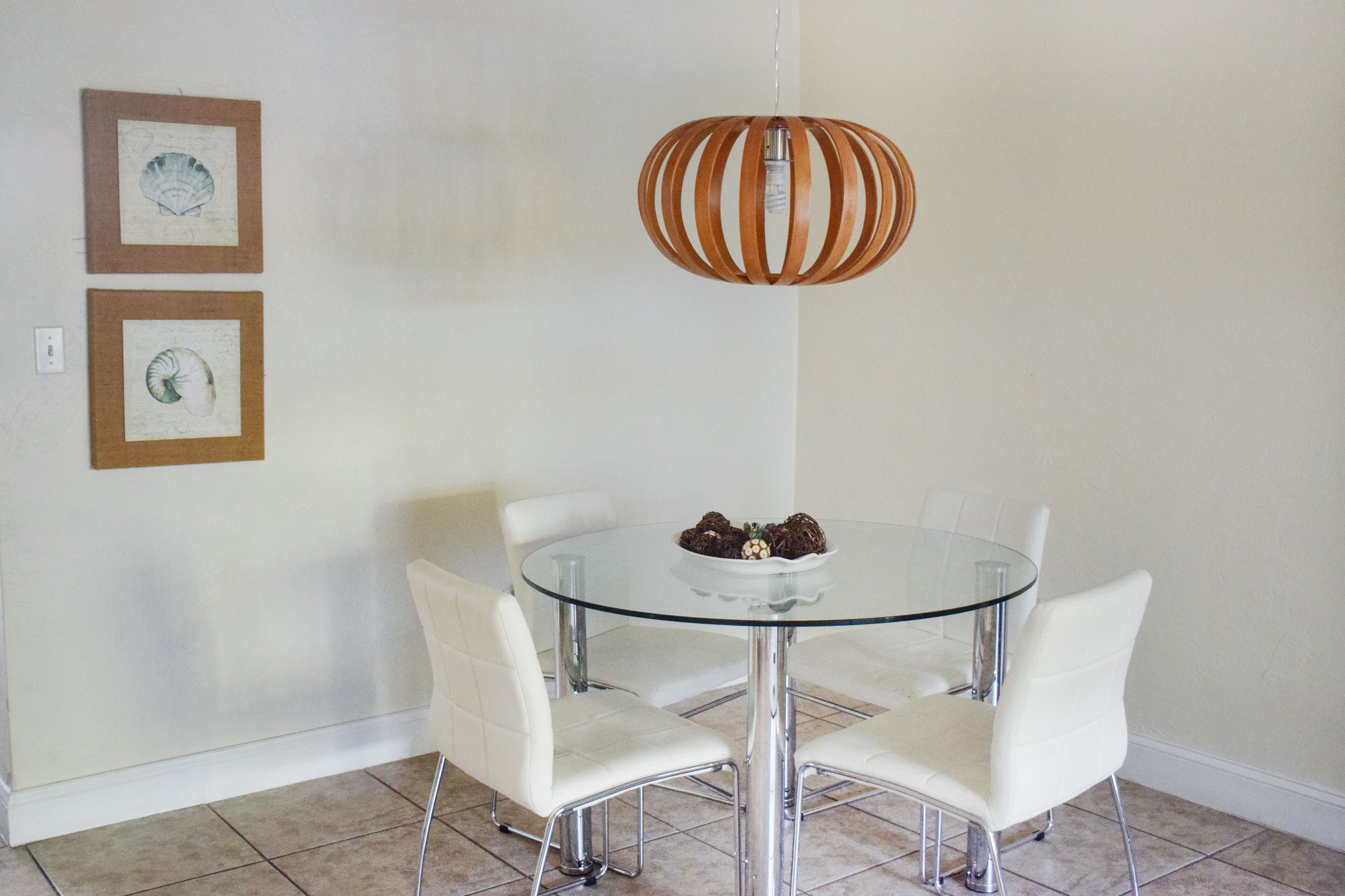 Un coin repas avec une table en verre et des chaises blanches, un luminaire suspendu en bois et deux tableaux encadrés au mur.