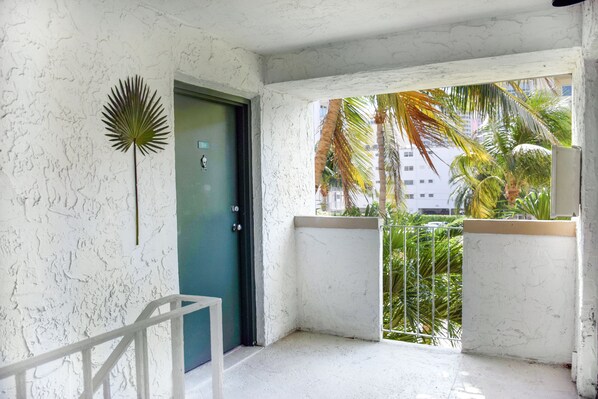 Una habitación con pared blanca texturizada, una puerta verde y vistas a palmeras y edificios desde afuera.