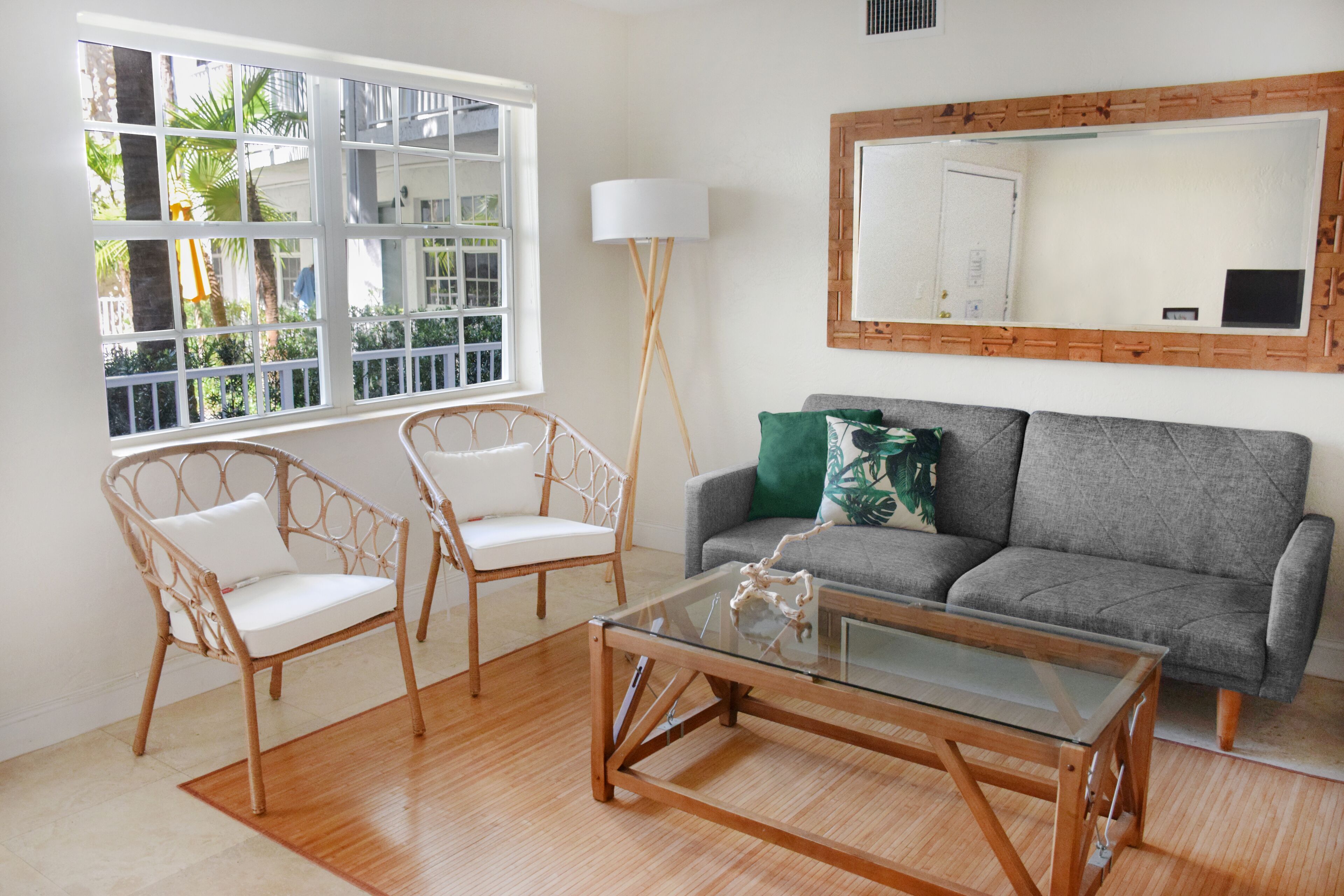 A living room with a glass coffee table, two white wicker chairs, a grey sofa with a green pillow, a floor lamp, and a large mirror on the wall.