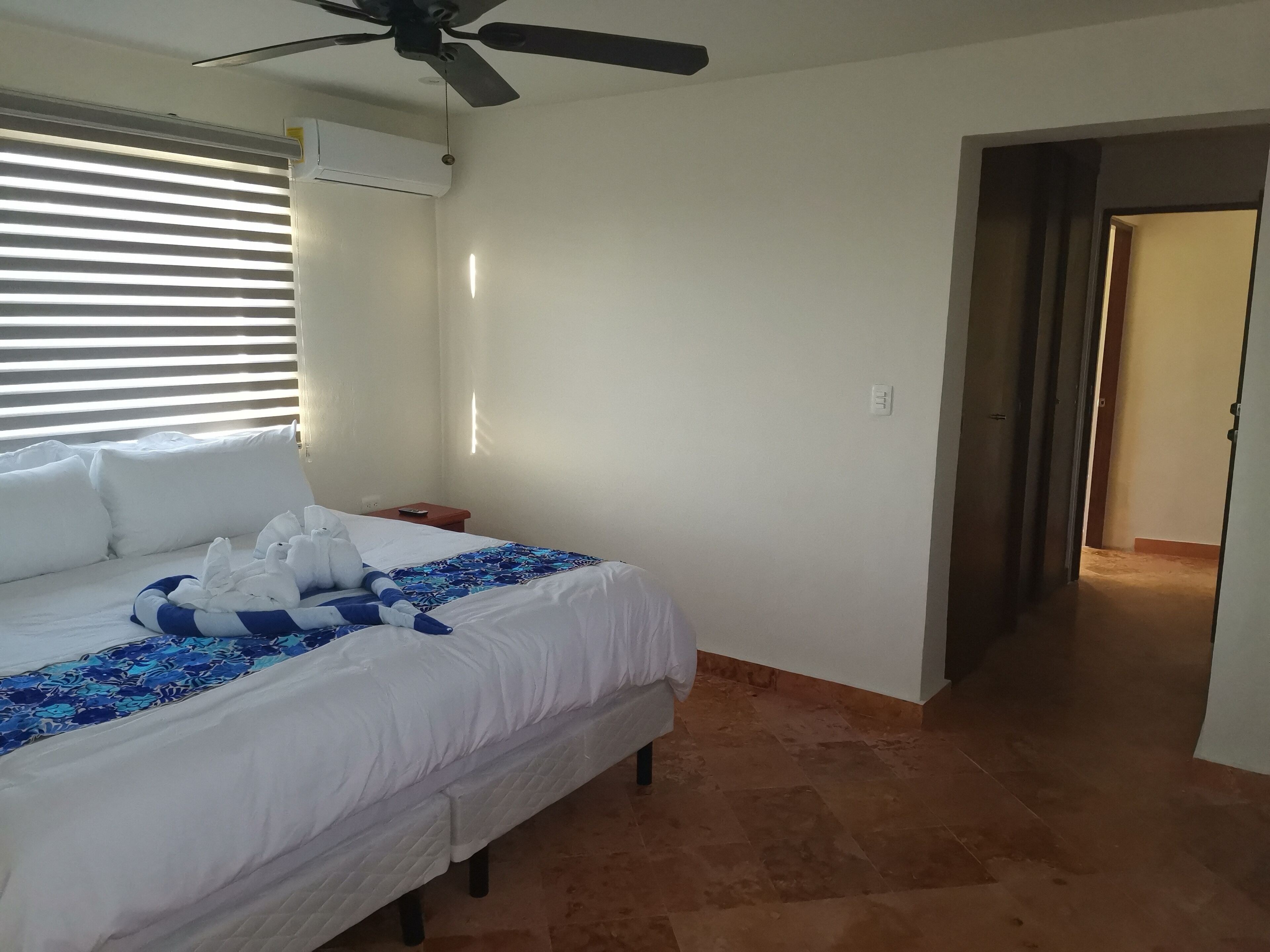 A hotel room with a bed, a ceiling fan, an air conditioning unit, a nightstand, and a hallway leading to another room.