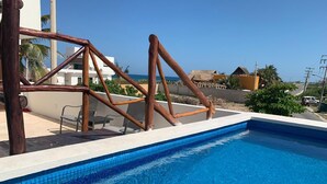 A swimming pool with a wooden deck structure, lounge chairs, and a view of the sea and buildings in the background. - Penthouse - Casa Mango Great location! (Isla Mujeres)