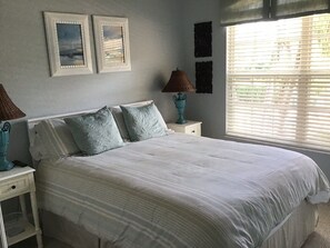 A bedroom with a bed, two bedside lamps, a window with blinds, and two framed pictures on the wall. - Beautiful Condo in Tranquil, Tropical Setting in Flamingo Fairways 2 (Naples)