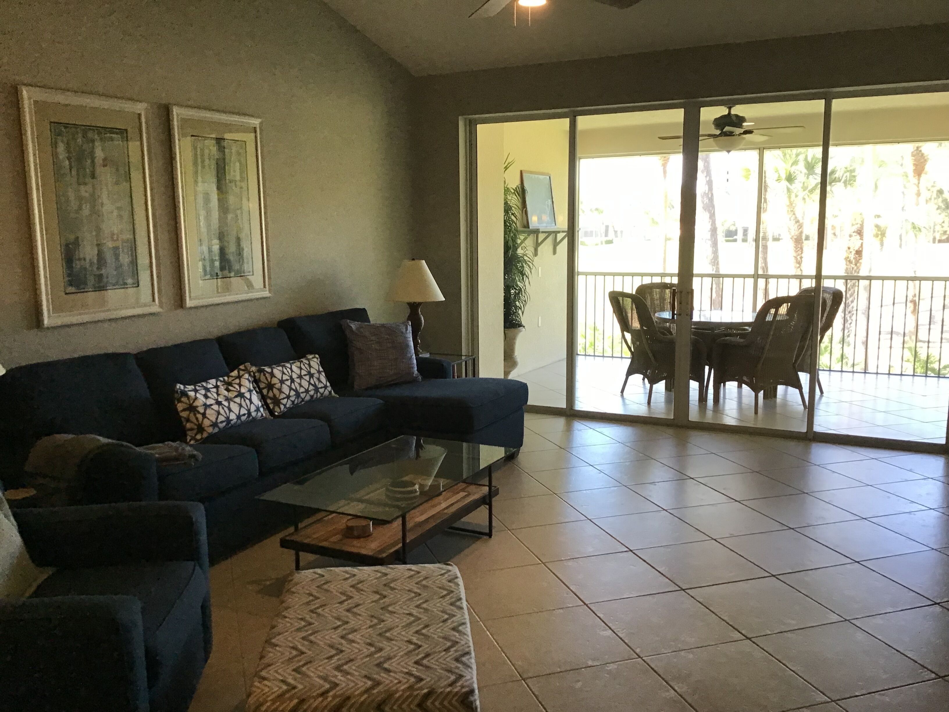 A living room with a dark blue sofa, a glass coffee table, and a patterned ottoman. There are two framed pictures on the wall, a lamp, and a sliding glass door leading to an outdoor patio with wicker furniture.