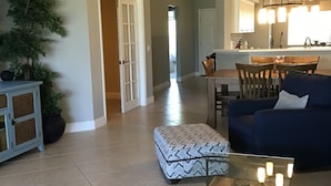 A spacious living room with a blue sofa, a glass coffee table, and a patterned ottoman. A dining area with a wooden table and chairs is visible in the background. - Beautiful Condo in Tranquil, Tropical Setting in Flamingo Fairways 2 (Naples)