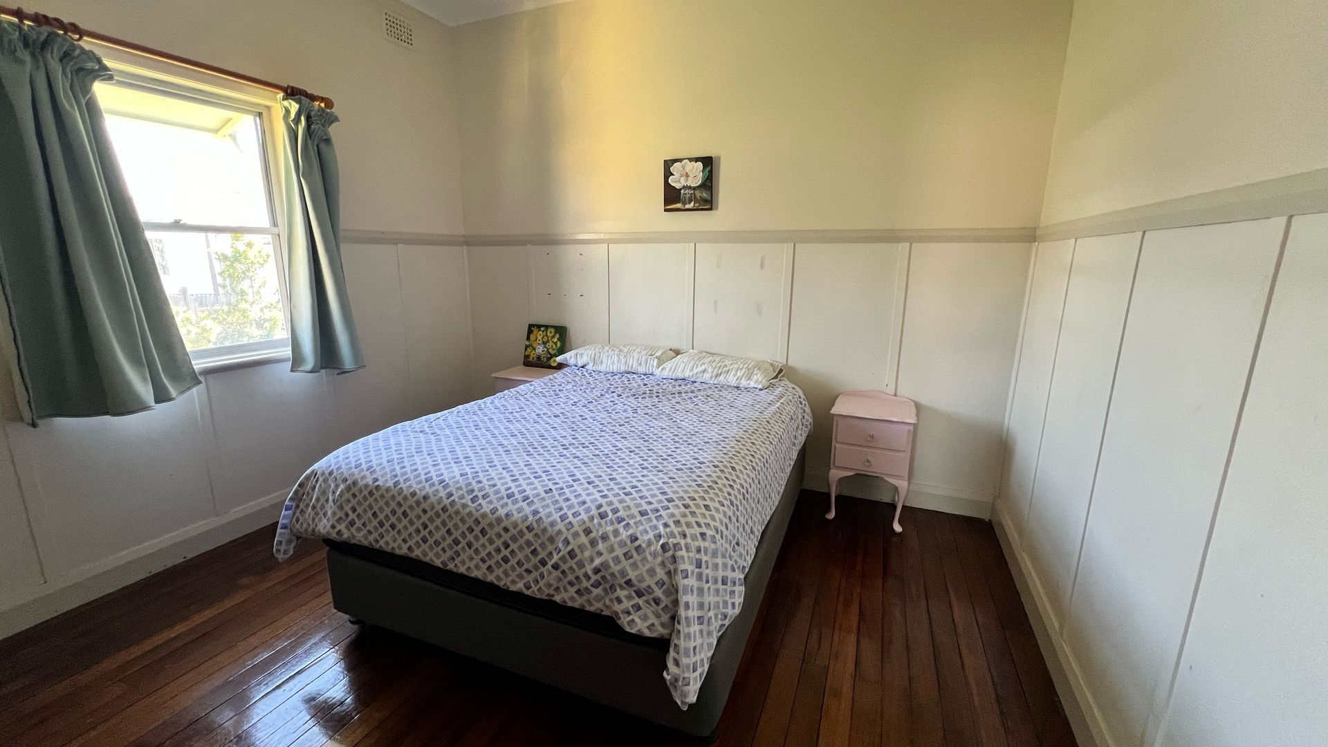 Une chambre à coucher avec un lit, une table de chevet, une fenêtre avec des rideaux et un tableau accroché au mur.