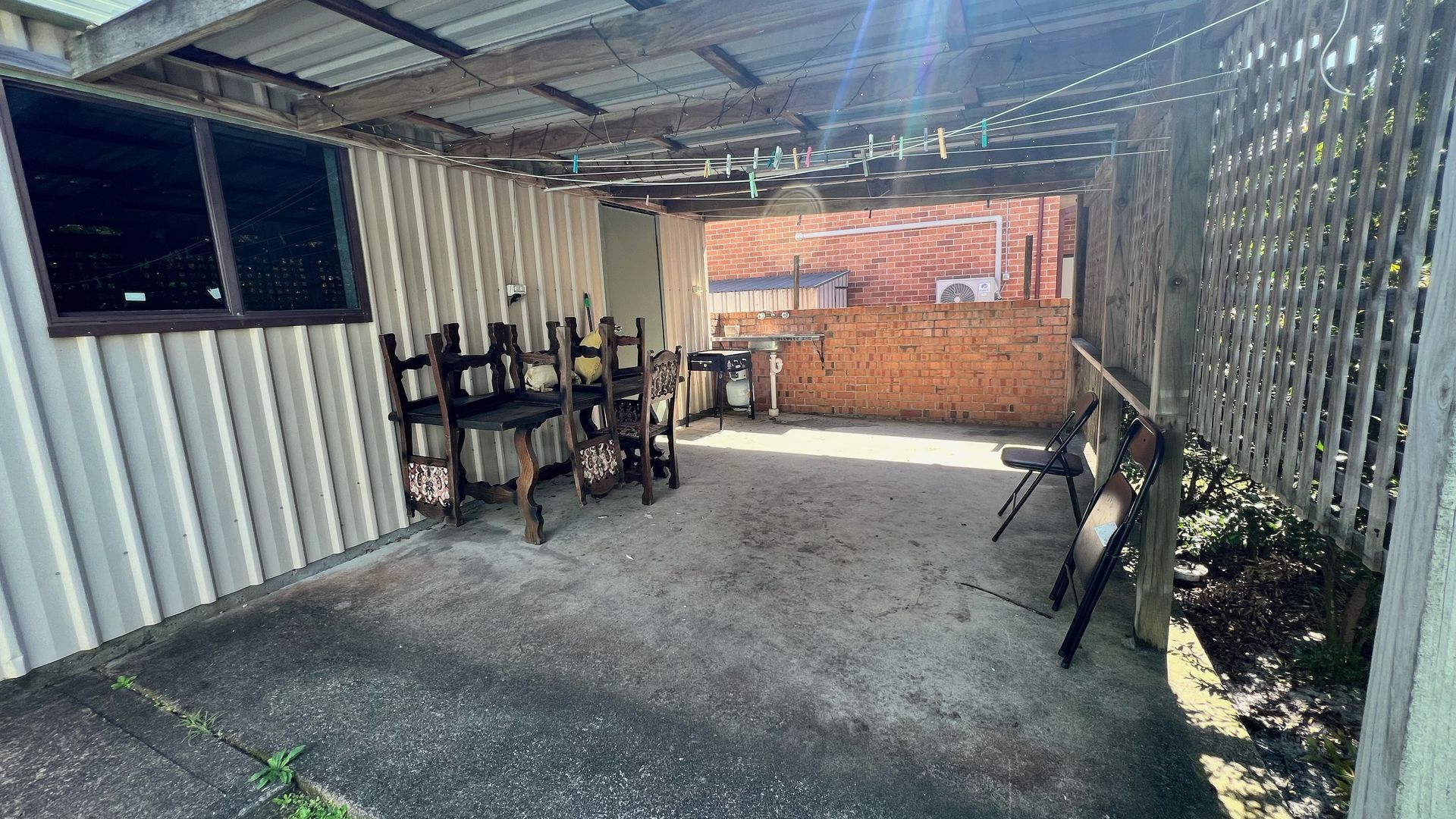 A covered outdoor area with a concrete floor, metal walls, and a brick wall in the background. There are several wooden chairs and a table.