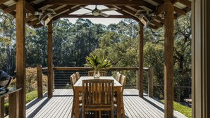 Um deck de madeira com mesa e cadeiras, ladeado por uma pérgula de madeira, com vista para uma área arborizada.