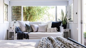 Um quarto aconchegante com um sofá branco, várias almofadas, um cobertor de tricô, um abajur e um vaso de planta.