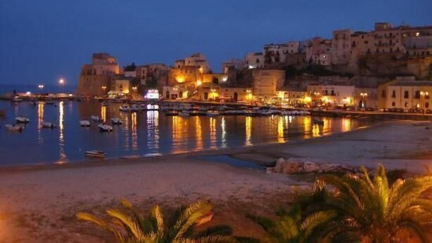 A coastal town at dusk with buildings along the waterfront, boats docked, and a sandy beach in the foreground.