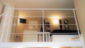 A room with a white bed, a desk, and a chair. There is a window with a view of the outside.