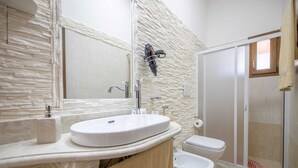 Une salle de bain avec un mur à l’aspect pierre, un lavabo blanc, des toilettes et un espace douche.