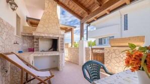 Un espace cuisine extérieur revêtu de pierres, comprenant un barbecue, un fauteuil de relaxation et une table recouverte d’une nappe à motifs floraux.